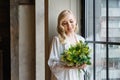 Wedding morning of the bride. Exquisite blonde in a delicate peignoir standing near the window with a bouquet of flowers in her Royalty Free Stock Photo