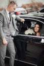 . The bride with a bouquet of roses sits smiling in a black car, the groom stands leaning on the car Royalty Free Stock Photo