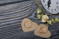 Wedding rings lie on hearts cut out of paper. Nearby a dried rose and a clock. On brushed pine boards painted in black and white