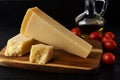 Wedge of italian hard cheese Grana Padano or Parmesan on a brown wood cutting boad, small red tomatoes and olive oil over black