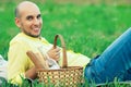 Weekend picnic concept. Portrait of a young handsome bald man in trendy clothes lying on green grass in the park. Picnic basket