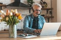 Weekend work in the home kitchen. Senior man in headset working on laptop. Additional earnings in retirement Royalty Free Stock Photo