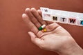 A weekly medicine dispenser opened for Sunday, hand holding pills and vitamins from white pill box on terracotta