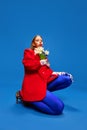 Weird girl in blue tights posing with flowers in unusual, strange, red jacket sitting against blue color studio Royalty Free Stock Photo