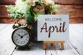 Welcome April text written on paper card with wooden easel and alarm clock with flower in metal vase decoration