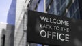 Welcome back to the Office on a black city-center sign in front of a modern office building