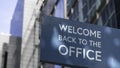 Welcome back to the Office on a blue city-center sign in front of a modern office building