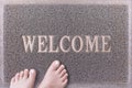 Welcome Door Mat With Female Feet. Friendly Grey Door Mat Closeup with Bare Woman Feet Standing. Welcome Carpet. Royalty Free Stock Photo