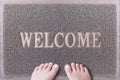 Welcome Door Mat With Female Feet. Friendly Grey Door Mat Closeup with Bare Woman Feet Standing. Welcome Carpet. Royalty Free Stock Photo