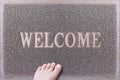 Welcome Door Mat With Female Foot. Friendly Grey Door Mat Closeup with Bare Woman Foot Standing. Welcome Carpet.