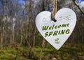 Welcome Spring greeting card with decorative white wooden heart with text on a blurred spring forest background.