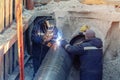 Welder welding water or gas steel pipeline with assistant worker in trench. City underground utilitites renewal and replacemen