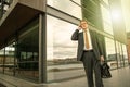 Well dressed businessman with briefcase standing talking on the phone in front of modern glass building  with sun reflection. Pers Royalty Free Stock Photo