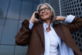 well-groomed healthy slender gray-haired mature business lady dressed in a stylish brown jacket uses a smartphone to