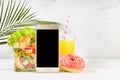 Wellness summer food set of orange juice, pink donut, tropical shrimp salad in box, blank phone in white bar interior, palm leaf. Royalty Free Stock Photo