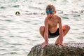 A boy with glasses for swimming sits on a large rock in the sea