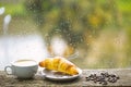 Wet glass window and cup of hot caffeine beverage. Coffee drink with croissant dessert. Enjoying coffee on rainy day Royalty Free Stock Photo