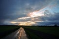 Wet reflecting country road made of concrete slabs is leading through the dark fields under a dramatic cloudy sky with evening sun Royalty Free Stock Photo