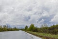 Wet rural road, going into the distance. Around dense thickets of trees and shrubs. Cloudy sky