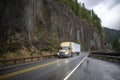 Wet yellow big rig semi truck transporting cargo in dry van semi trailer running on the mountain winding slippery road with rock Royalty Free Stock Photo