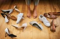 What shall I wear. an unrecognizable womans feet seen next to a wide variety of womans shoes on the floor at home.