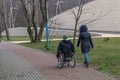 Wheelchair user in black with backpack on back with girlfriend in black jacket and jeans on walk along tiled sidewalk among trees