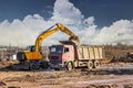 A wheeled excavator loads a dump truck with soil and sand. An excavator with a high-raised bucket against a cloudy sky View from Royalty Free Stock Photo