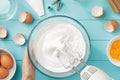 Whipped egg whites cream to perfect peaks in glass bowl with mixer and hand on blue wooden table. Royalty Free Stock Photo