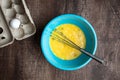 Whipped eggs in blue mixing bowl, metal whisk, whole egg in cardboard carton, wood table