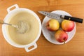 Whisk in white pan with dough, apples, kitchen knife in plate on wooden table. Top view