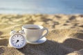 A white alarm clock and a cup of tea or coffee standing on the sand against the background of the blue sea at sunrise. Breakfast Royalty Free Stock Photo