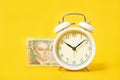 White alarm clock and money on a yellow background. Ukrainian hryvnia.