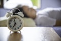 A white alarm clock rests on a table with a woman lying down and