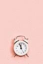 White alarm clock shows about twelve oclock on pink paper background covered with sequins with copy space