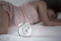 alarm clock on white bed with blured image of woman in pink sleepwear