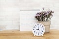 White analog clock with blurred white calendar and purple flower in ceramic vase on wooden desk