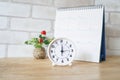 White analog clock with blurred calendar on wooden desk