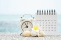 White analog clock and opened calendar with blurred shell and plumeria flower on blurred sea background