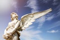 White angel marble sculpture with open long wings across the frame and against a bright sunny blue sky with lens flare