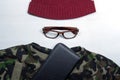 On a white background lies a camouflage men`s t-shirt, brown glasses and a red men`s hat