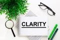 On a white background lies a notebook with the word CLARITY, glasses, a magnifying glass, green markers and a green plant