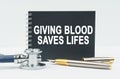 On a white background lies a stethoscope, a pen and a black notebook with the inscription - GIVING BLOOD SAVES LIFES Royalty Free Stock Photo