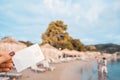 White Bank Card In Woman Hand On Background Of Hotel Beach, Family Comes Out Of Sea, Moraitika, Corfu, Greece. The Concept Of