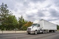 White big rig day cab semi truck with roof spoiler transporting goods in dry van semi trailer driving on the multiline highway