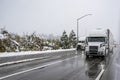 White big rig semi trucks with turned on headlight transporting cargo in semi trailer running on the winter highway road with wet Royalty Free Stock Photo