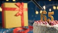 White birthday cake number 65 golden candles burning by lighter, blue background with lights and gift yellow box tied up with red Royalty Free Stock Photo