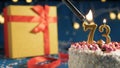 White birthday cake number 73 golden candles burning by lighter, blue background with lights and gift yellow box tied up with red Royalty Free Stock Photo