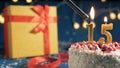 White birthday cake number 15 golden candles burning by lighter, blue background with lights and gift yellow box tied up with red Royalty Free Stock Photo