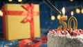 White birthday cake number 50 golden candles burning by lighter, blue background with lights and gift yellow box tied up with red Royalty Free Stock Photo