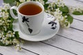 White and black cup of black or herbal tea on saucer with white bird cherry tree flowers on white wooden table